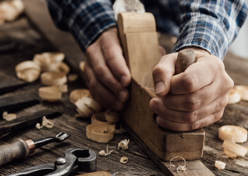 A carpenter carefully shapes wood using a hand plane to create a pleasing and high-quality display piece. Other tools for hand-carving fine wood products are visible.